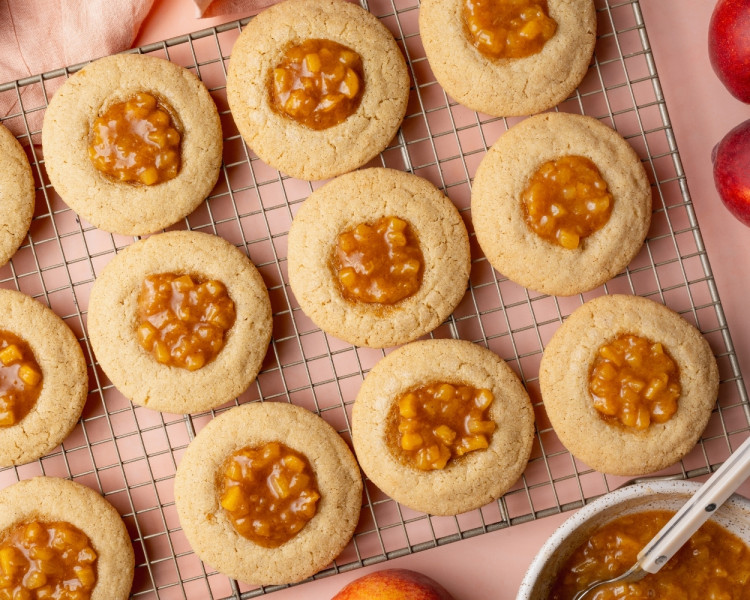 Overhead image of peach cobbler cookies cooling on a wire rack with homemade peach filling spooned into the centers. Fresh peaches and a bowl of peach filling are styled around the cookies on a blush pink background.