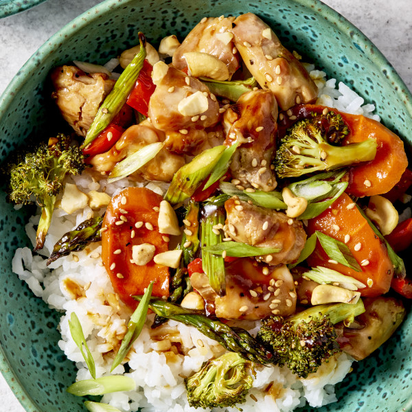 Sheet pan sesame chicken and veggies served in a bowl with on top of rice and garnished with green onions and sesame seeds.