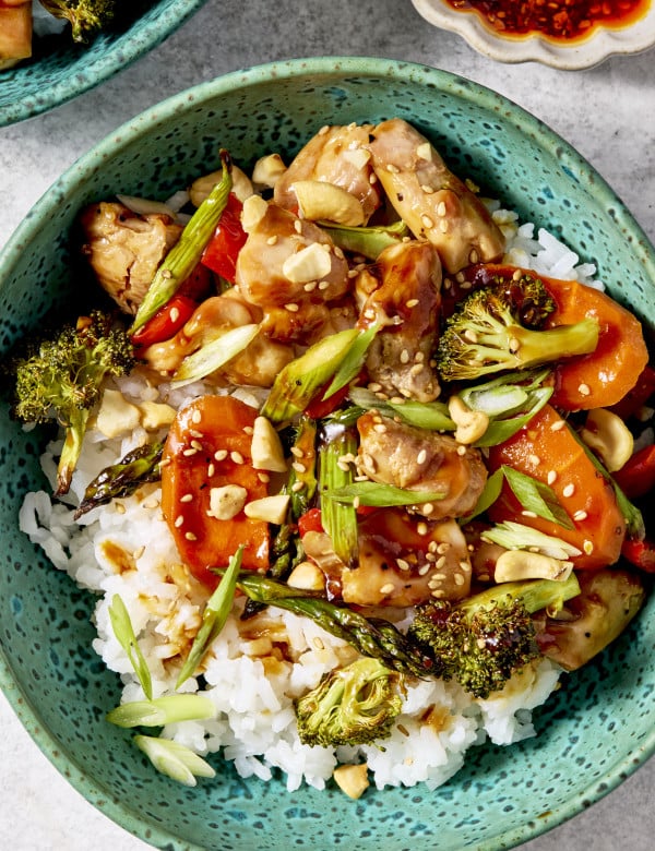 Sheet pan sesame chicken and veggies served in a bowl with on top of rice and garnished with green onions and sesame seeds.