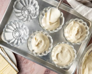 Close-up of a glass dessert cup filled with white chocolate mousse topped with whipped cream, fresh raspberries, and white chocolate curls, with more mousse cups softly blurred in the background.
