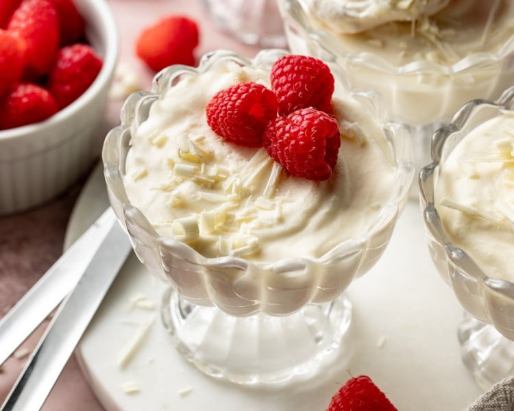 Overhead close-up of a spoon scooping fluffy white chocolate mousse from a glass dessert cup topped with raspberries and white chocolate shavings. Additional mousse cups and fresh raspberries surround the dessert.