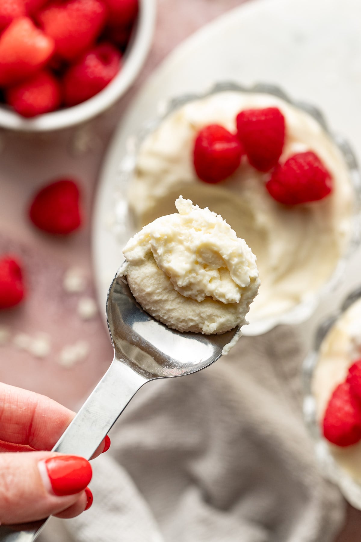 Close-up of a spoon holding a bite of white chocolate mousse topped with whipped cream. A hand with red nail polish holds the spoon, while blurred mousse cups and raspberries appear in the background.