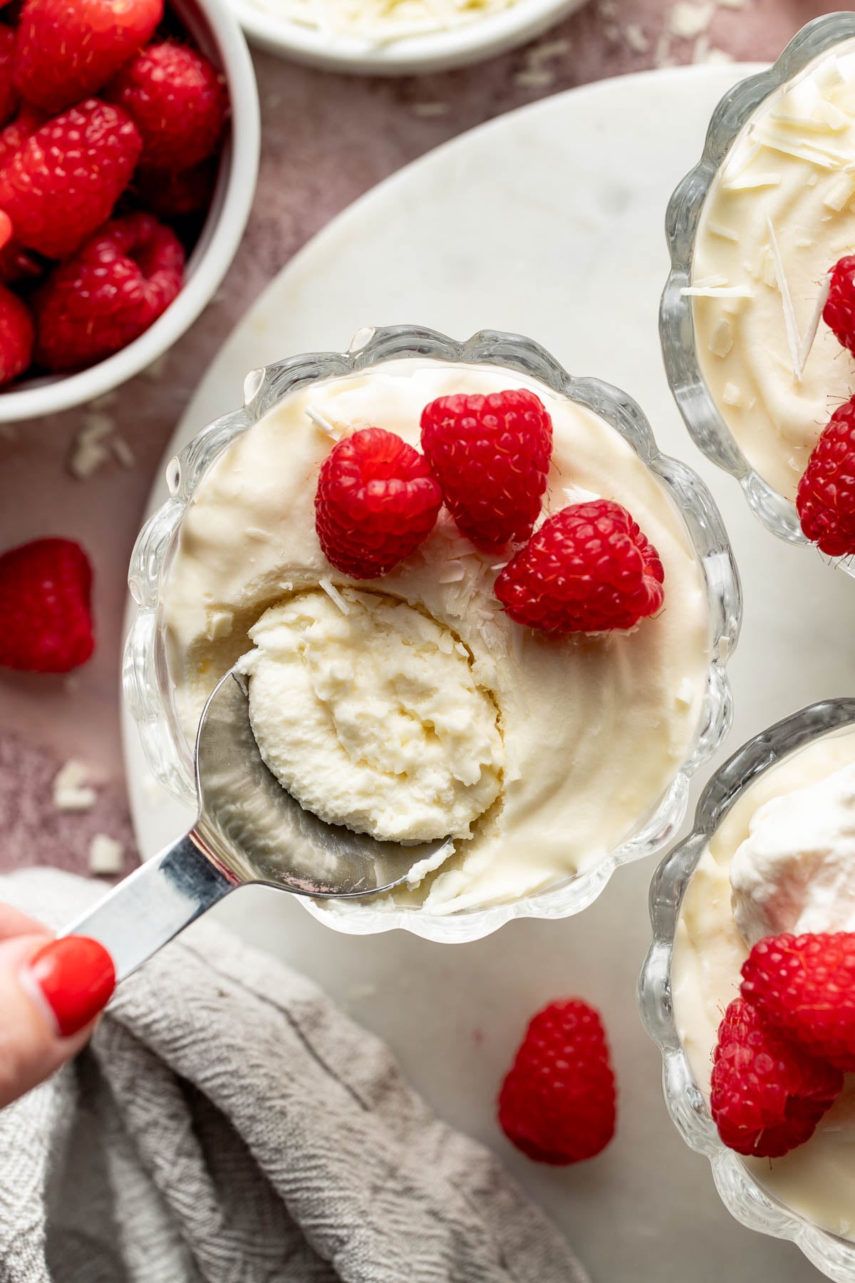 Overhead close-up of a spoon scooping creamy white chocolate mousse from a glass dessert cup topped with raspberries and white chocolate curls. Additional mousse cups and fresh raspberries surround the scene.