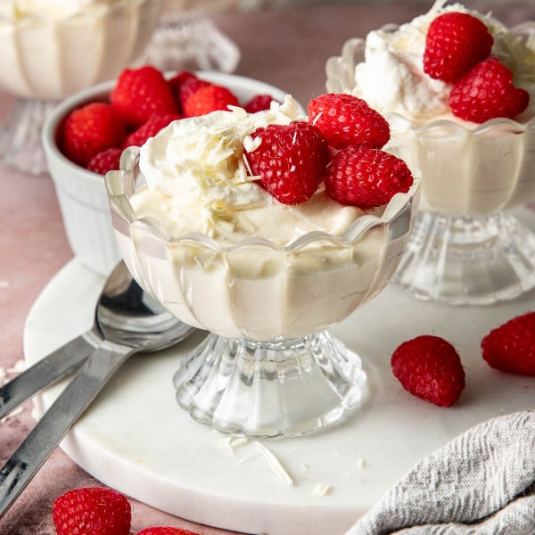 Angled hero shot of white chocolate mousse served in scalloped glass dessert cups topped with whipped cream, raspberries, and white chocolate shavings. Fresh raspberries, dessert spoons, and a linen towel are styled around the desserts on a pink tabletop.