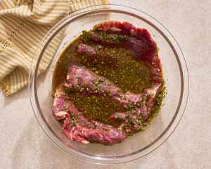 Flank steak in a glass mixing bowl with marinade poured over it.