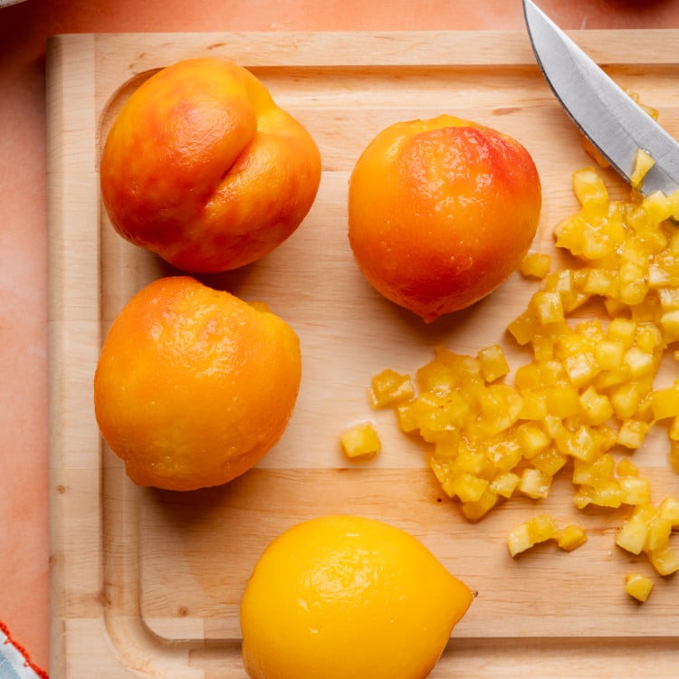 Peeled peaches on a cutting board with one of the peaches diced into small squares.