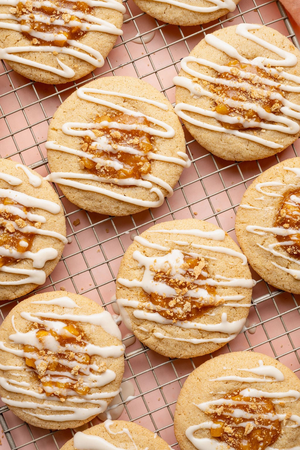 Overhead image of peach cobbler cookies arranged on a wire cooling rack over a blush pink background. The cookies are topped with peach cobbler filling, white glaze drizzle, and crushed graham cracker crumbs.