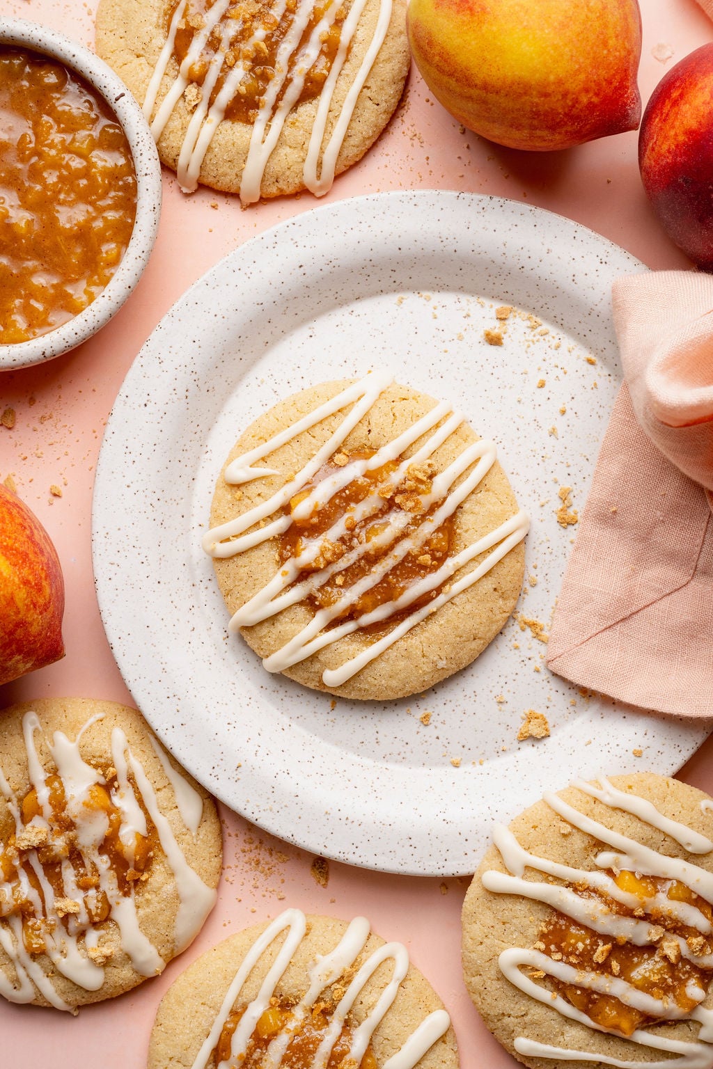 Overhead styled image featuring a peach cobbler cookie on a speckled ceramic plate surrounded by fresh peaches, a bowl of peach filling, and more glazed cookies on a blush pink background.