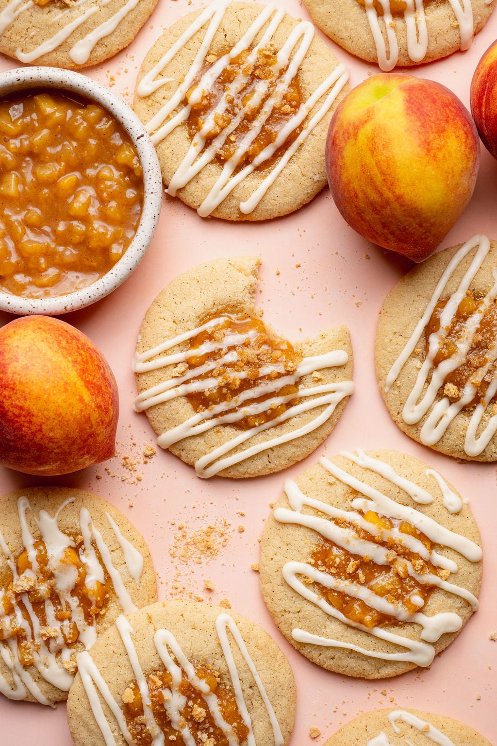 Overhead image of peach cobbler cookies styled with fresh peaches and a bowl of homemade peach filling on a blush pink background. One cookie has a bite taken out to reveal the soft interior beneath the peach topping and glaze drizzle.