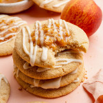 Close-up image of a stack of peach cobbler cookies drizzled with white glaze and topped with peach filling and graham cracker crumbs. The top cookie has a bite taken out, showing the soft and chewy texture inside, with fresh peaches and more cookies blurred in the background.
