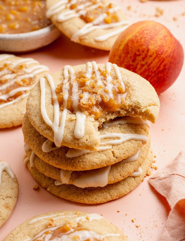 Close-up image of a stack of peach cobbler cookies drizzled with white glaze and topped with peach filling and graham cracker crumbs. The top cookie has a bite taken out, showing the soft and chewy texture inside, with fresh peaches and more cookies blurred in the background.