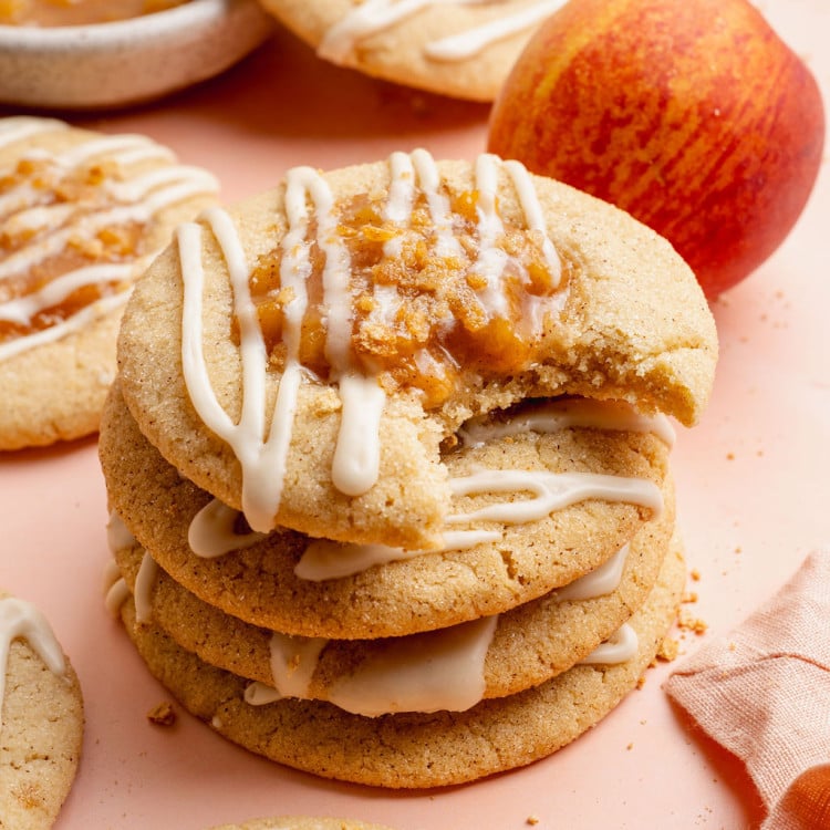 Close-up image of a stack of peach cobbler cookies drizzled with white glaze and topped with peach filling and graham cracker crumbs. The top cookie has a bite taken out, showing the soft and chewy texture inside, with fresh peaches and more cookies blurred in the background.