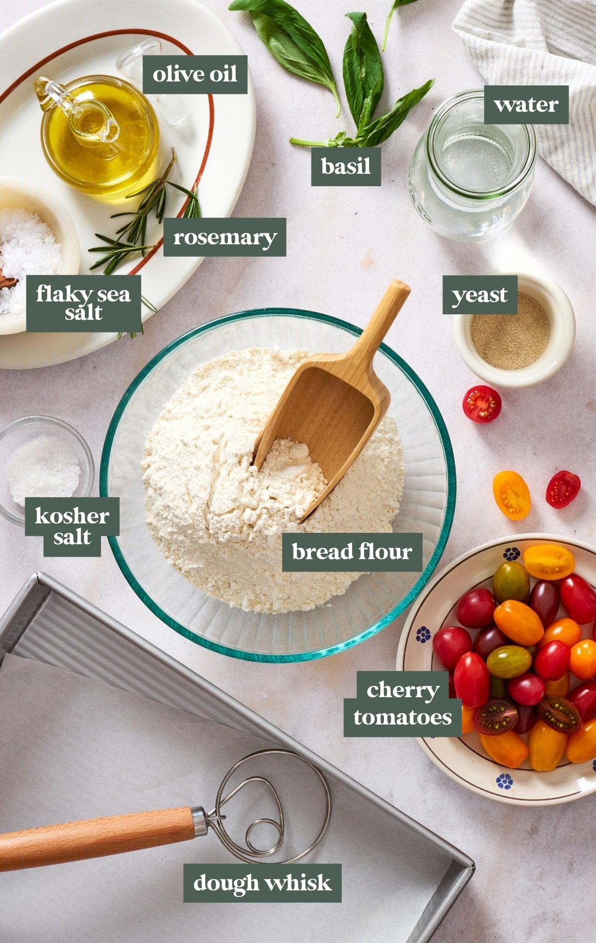Ingredients for homemade same day focaccia including bread flour, water, yeast, olive oil, kosher salt, flaky sea salt, rosemary, basil, cherry tomatoes, and a dough whisk.