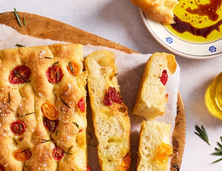 Finished homemade focaccia topped with cherry tomatoes, fresh rosemary, flaky sea salt, and olive oil, served with olive oil and balsamic vinegar for dipping.