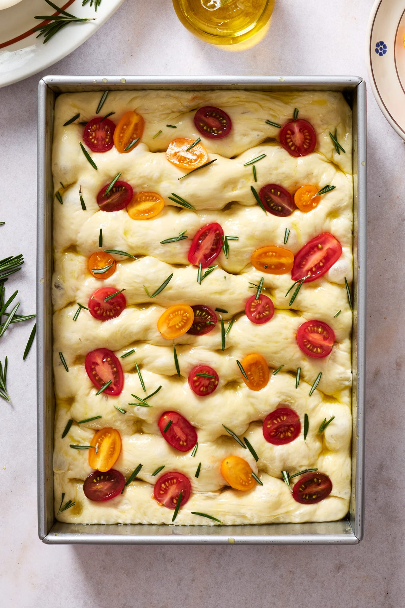 Unbaked same-day focaccia dough in a sheet pan after dimpling, topped with halved cherry tomatoes, fresh rosemary, and olive oil before baking.