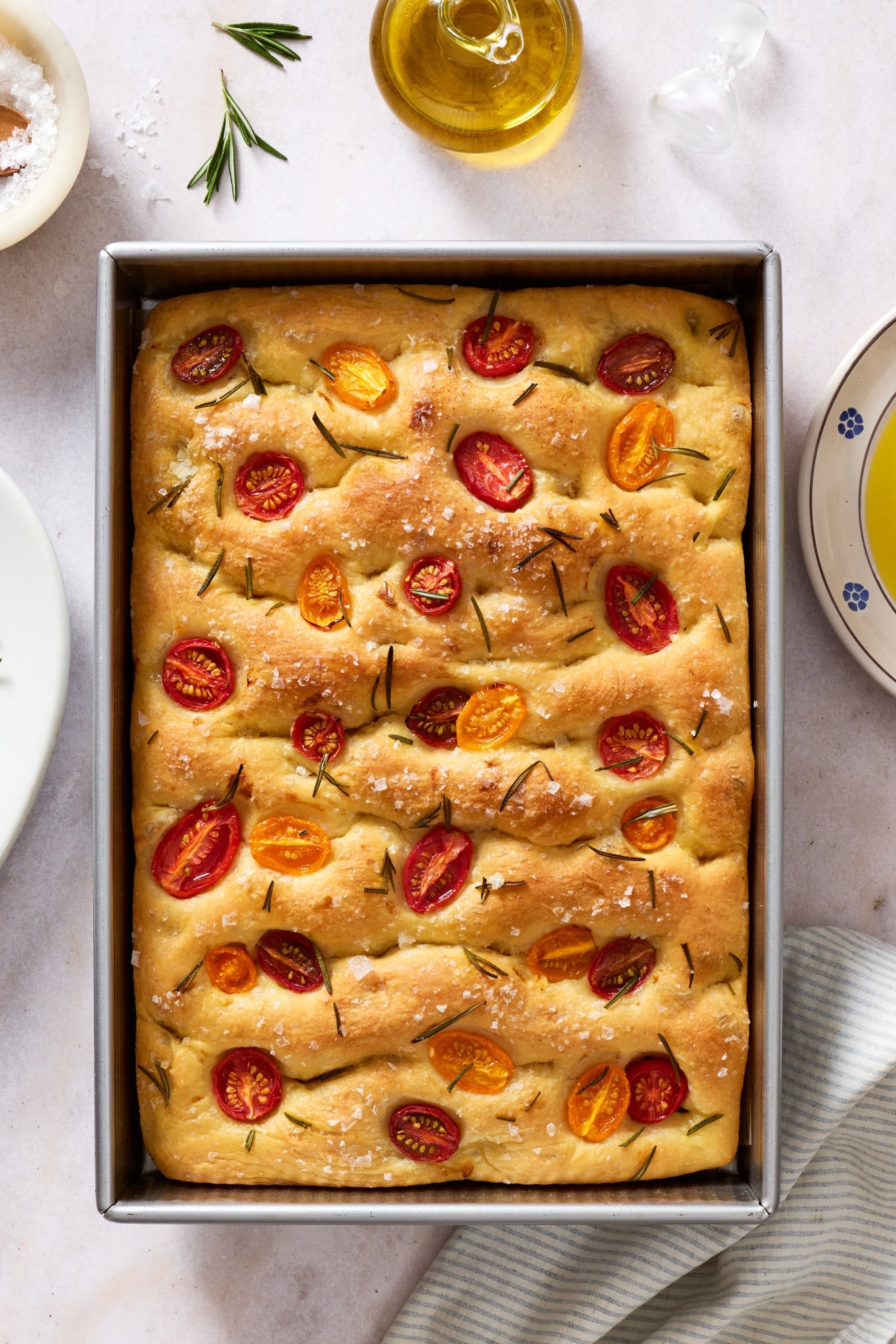 Golden brown tomato rosemary focaccia baked in a sheet pan and topped with cherry tomatoes, fresh rosemary, flaky sea salt, and olive oil.