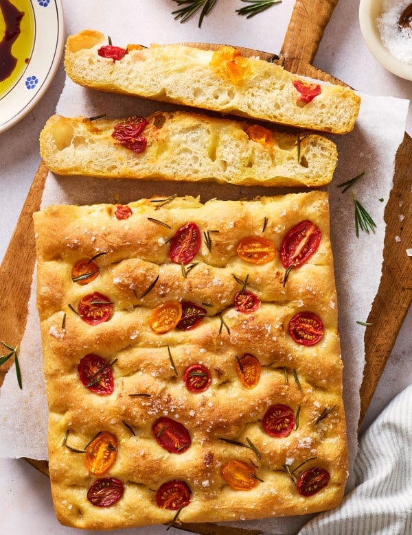 Whole tomato rosemary focaccia on a wooden board with sliced pieces showing the light, airy interior, served with olive oil and balsamic vinegar for dipping.