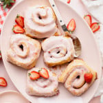 Five strawberry cinnamon rolls on a large oval plate with spoon and extra fresh strawberries.