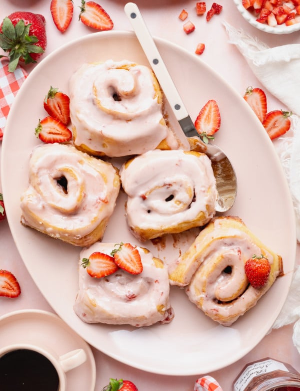 Five strawberry cinnamon rolls on a large oval plate with spoon and extra fresh strawberries.