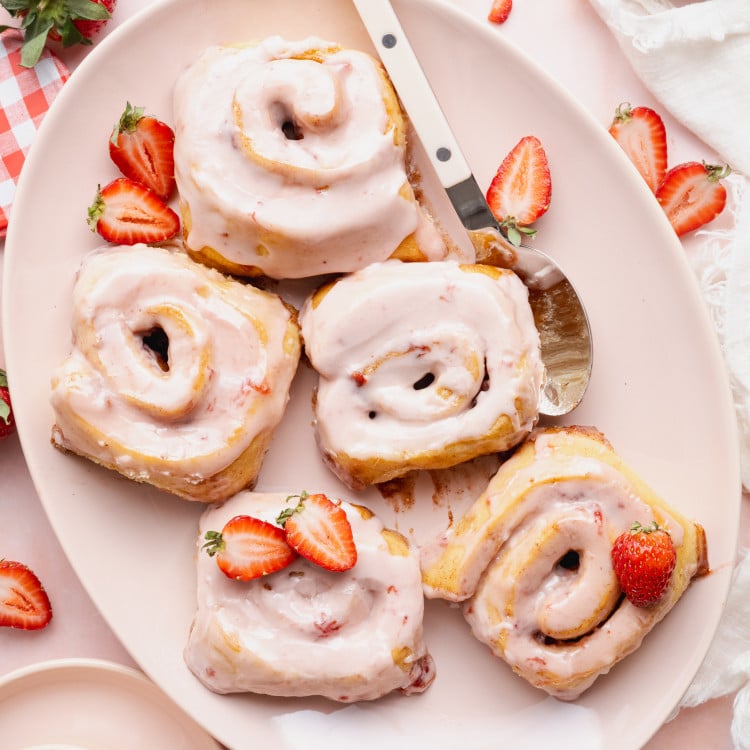 Five strawberry cinnamon rolls on a large oval plate with spoon and extra fresh strawberries.