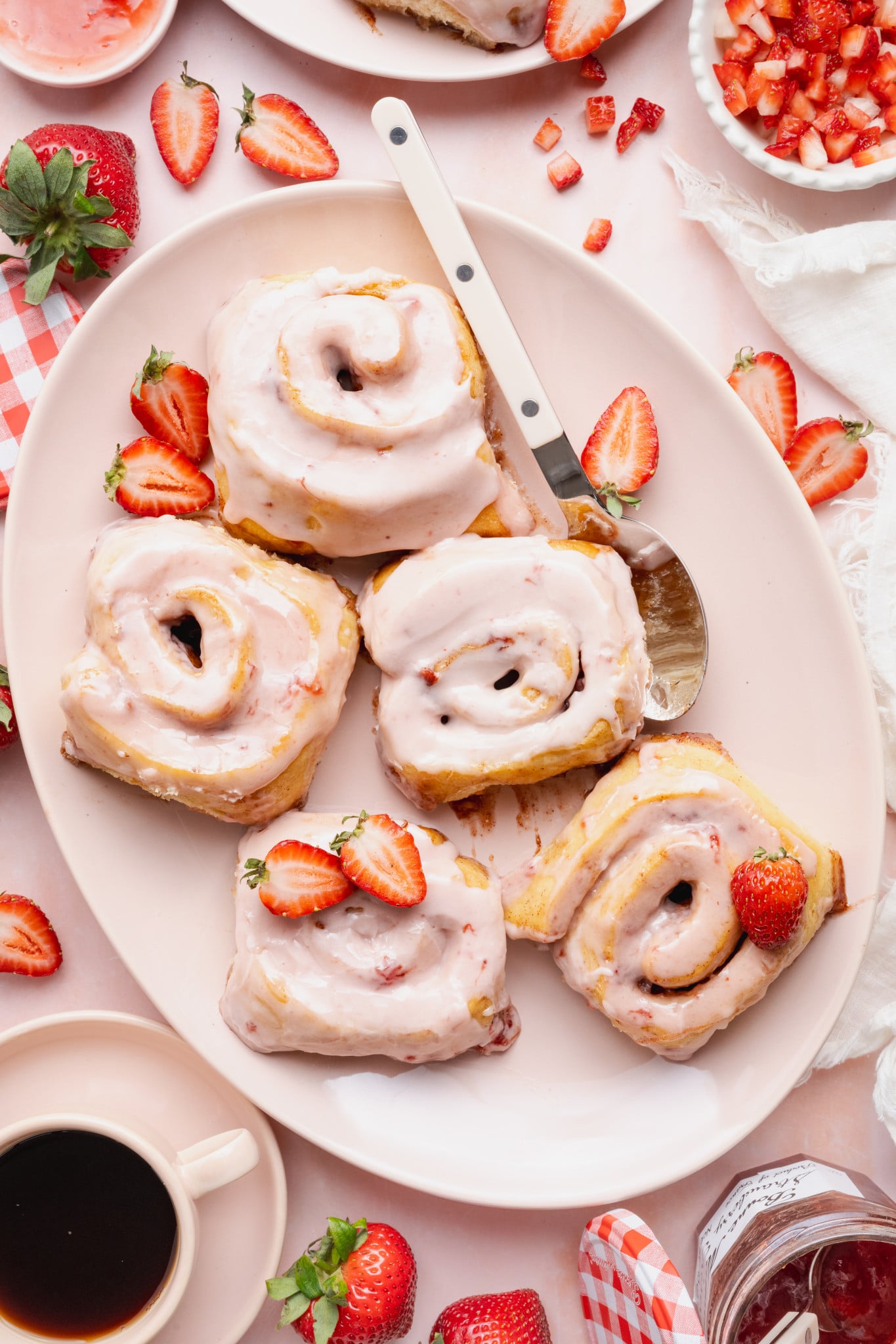 Five strawberry cinnamon rolls on a large oval plate with spoon and extra fresh strawberries.