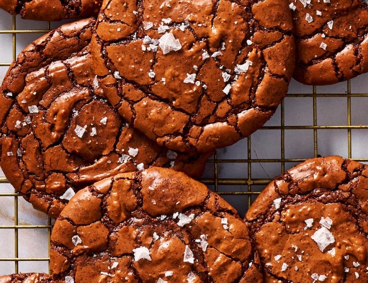 Crackly top brownie cookies piled on a cooling rack and sprinkled with flaky salt.