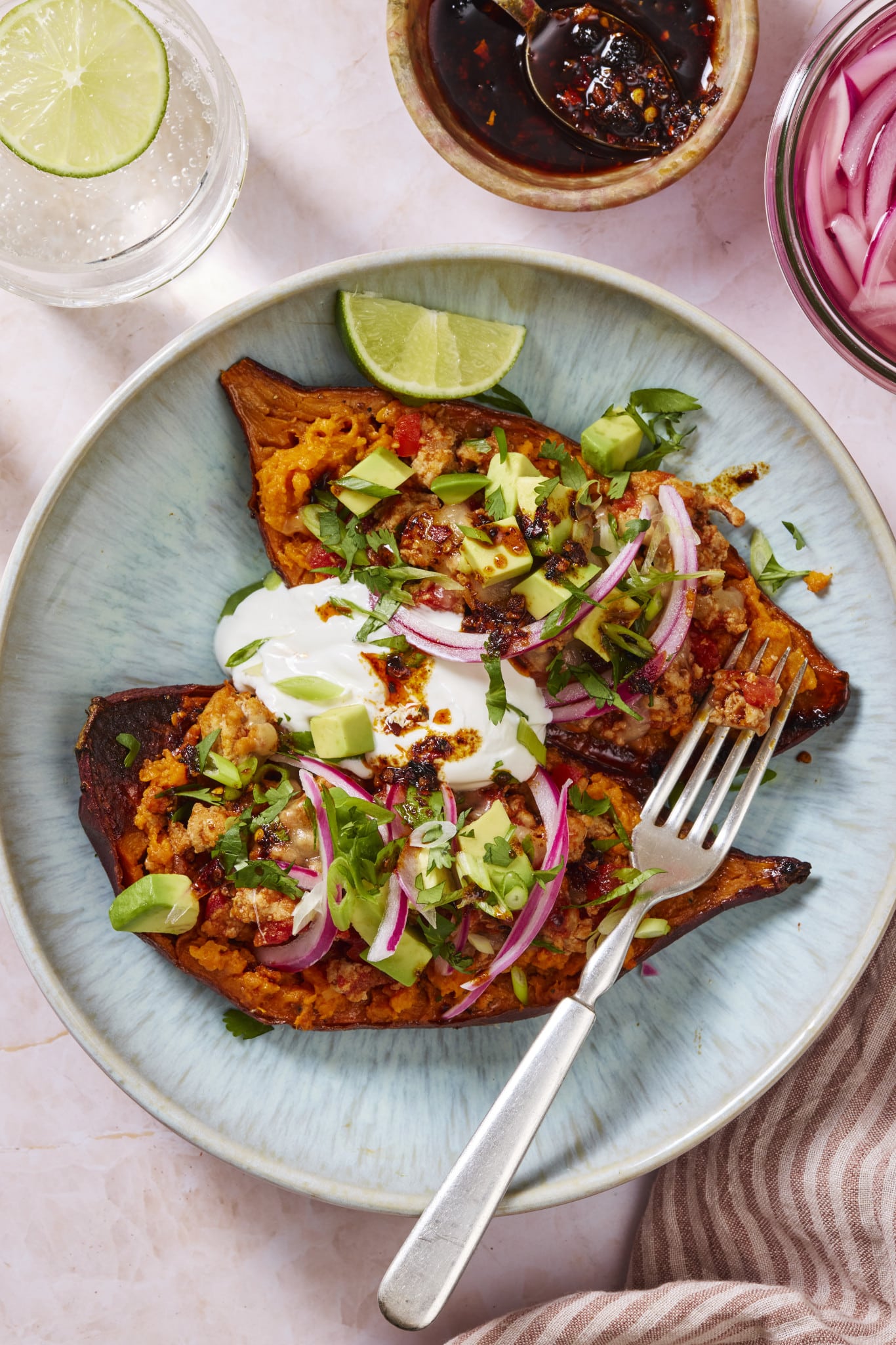 Two sweet potato halves topped with ground turkey, avocado, sour cream, pickled red onions and cilantro on a plate with a fork.