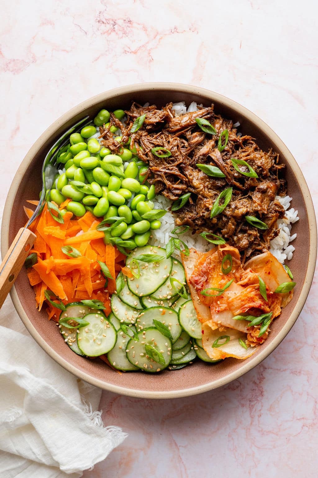 Slow cooker Korean-inspired beef bowl made with shredded beef, jasmine rice, edamame, shredded carrots, cucumber salad, kimchi, sesame seeds, and sliced green onions.