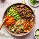 Overhead photo of a Korean-inspired beef bowl topped with tender shredded beef, edamame, cucumber salad, kimchi, shredded carrots, green onions, sesame seeds, and a drizzle of sriracha.