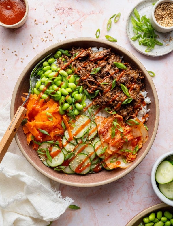 Overhead photo of a Korean-inspired beef bowl topped with tender shredded beef, edamame, cucumber salad, kimchi, shredded carrots, green onions, sesame seeds, and a drizzle of sriracha.