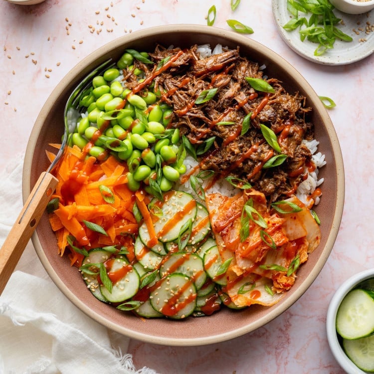 Overhead photo of a Korean-inspired beef bowl topped with tender shredded beef, edamame, cucumber salad, kimchi, shredded carrots, green onions, sesame seeds, and a drizzle of sriracha.