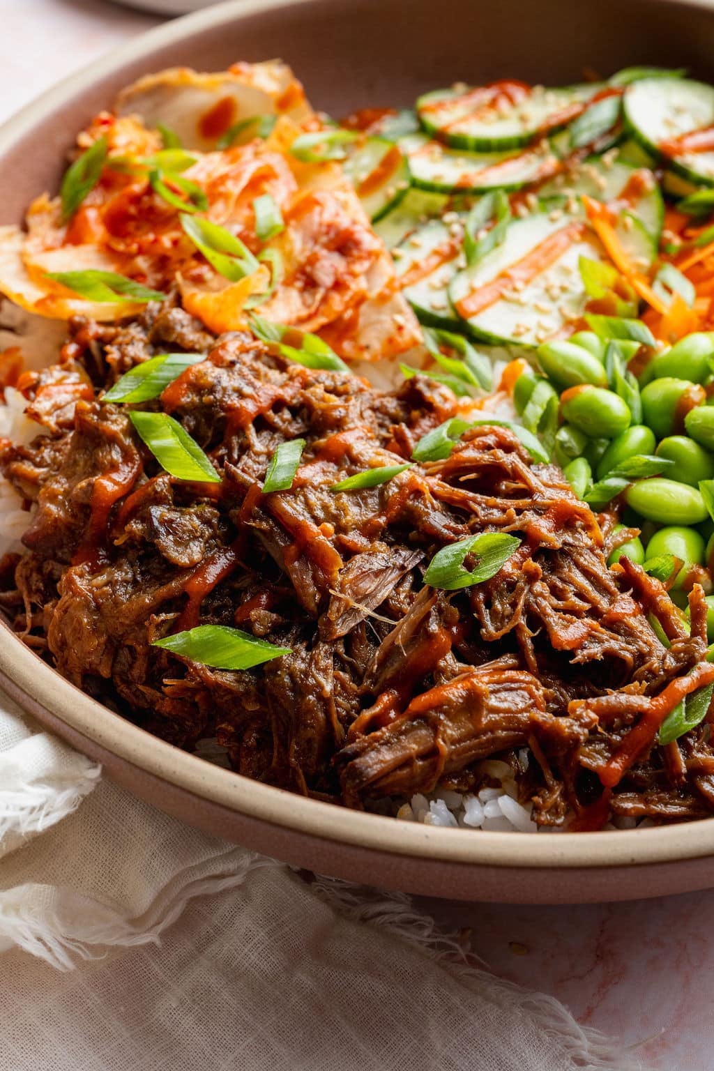 Close-up of tender slow cooker Korean-inspired shredded beef served over white rice with kimchi, cucumber salad, edamame, green onions, and sriracha.