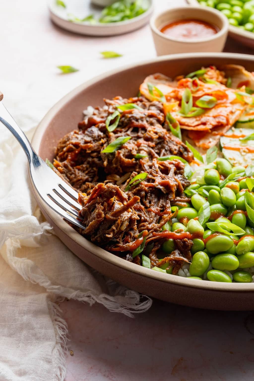 Fork lifting a bite of slow cooker Korean-inspired shredded beef from a rice bowl with edamame, kimchi, cucumber salad, carrots, and green onions.