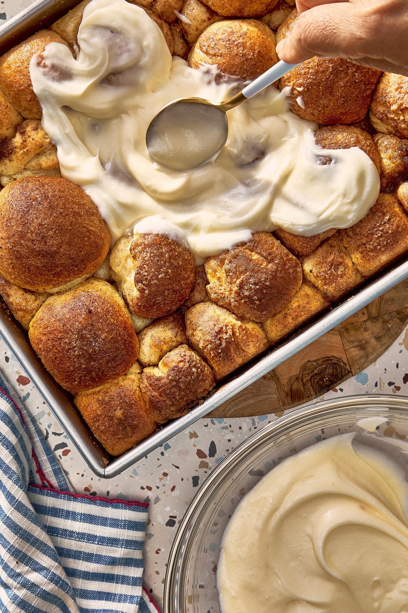 Cream cheese frosting being spread over warm, golden brown cinnamon roll bites in a baking pan.