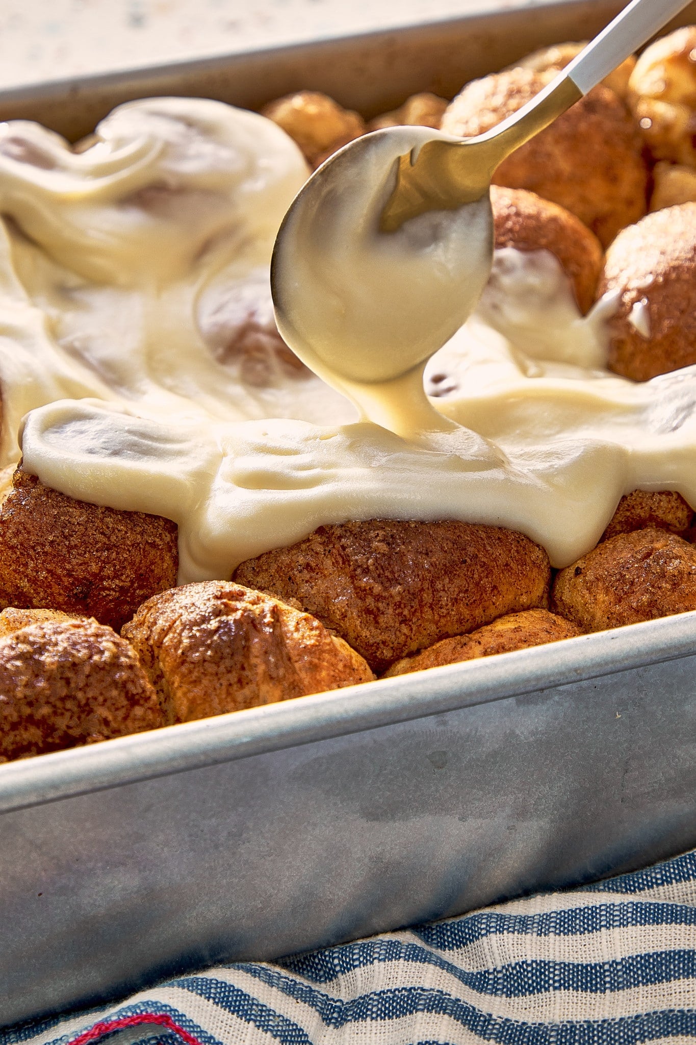 Thick cream cheese frosting dripping from a spoon as it is spread over freshly baked cinnamon roll bites.