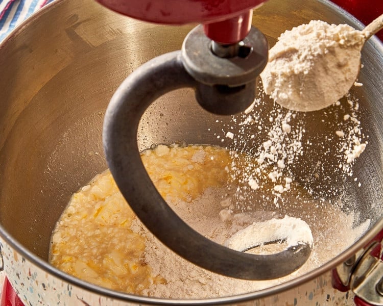 Bread flour being gradually added to the wet ingredients in a stand mixer fitted with a dough hook.