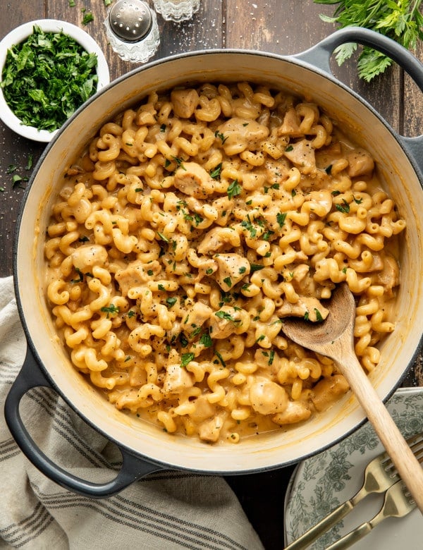 Chicken mac and cheese cooked in a dutch oven and garnished with fresh parsley with wood spoon on the bottom right side.