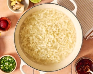 Wide noodles cooking in a large pot of boiling water with green onions, fresh ginger, lime, and chili crisp nearby.