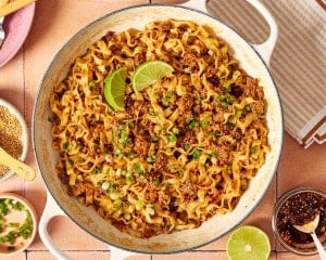 Finished saucy ground beef and noodles in a skillet, topped with green onions, sesame seeds, chili crisp, and fresh lime wedges.