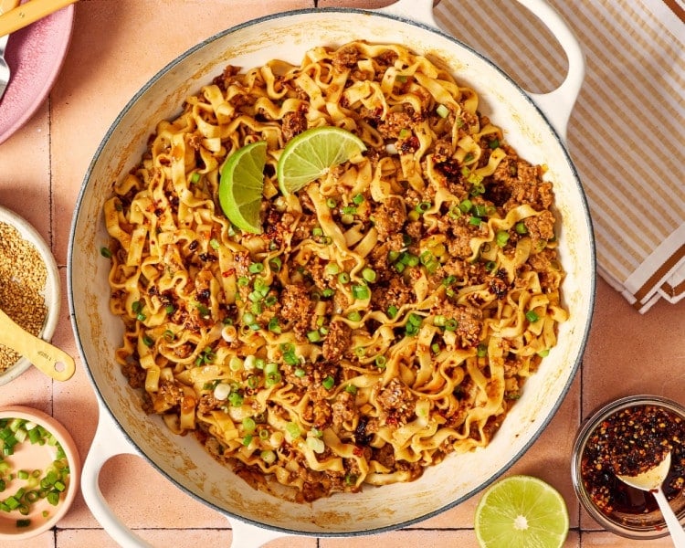 Finished saucy ground beef and noodles in a skillet, topped with green onions, sesame seeds, chili crisp, and fresh lime wedges.