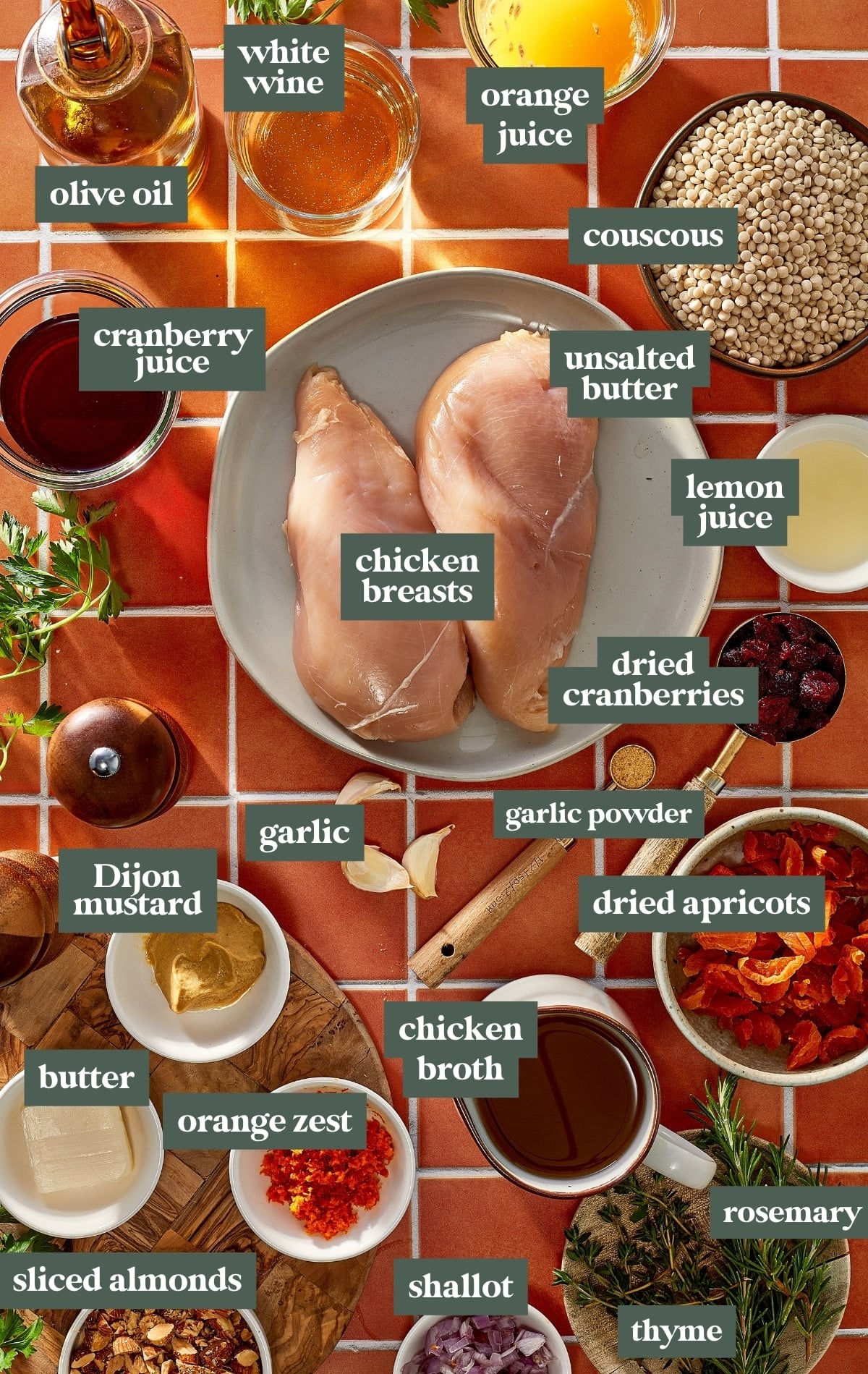 Overhead view of ingredients for cranberry apricot chicken, including chicken breasts, pearl couscous, dried cranberries, apricots, orange and cranberry juices, white wine, chicken broth, Dijon mustard, fresh herbs, and almonds.