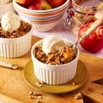 Two ramekins of apple crisp topped with vanilla ice cream with spoon digging into first ramekin.