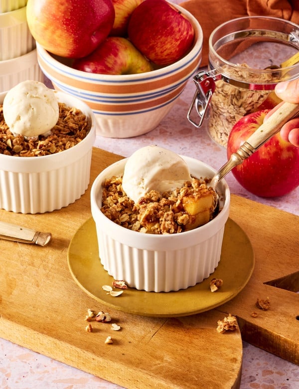 Two ramekins of apple crisp topped with vanilla ice cream with spoon digging into first ramekin.