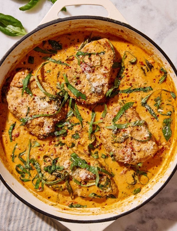 Overhead view of Marry Me Pork Chops nestled in a skillet of rich, creamy sun-dried tomato Parmesan sauce, garnished with fresh basil, grated Parmesan cheese, and red pepper flakes.