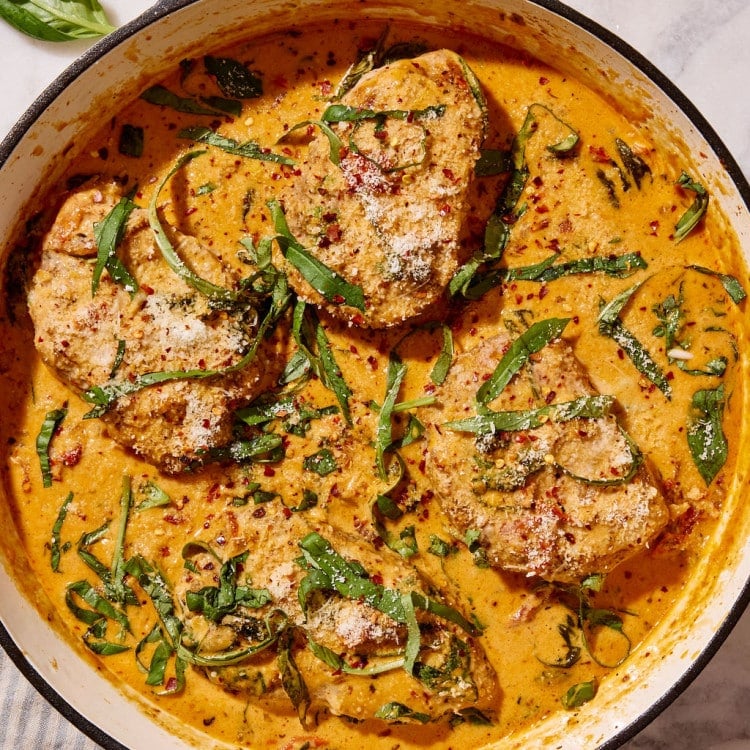 Overhead view of Marry Me Pork Chops nestled in a skillet of rich, creamy sun-dried tomato Parmesan sauce, garnished with fresh basil, grated Parmesan cheese, and red pepper flakes.