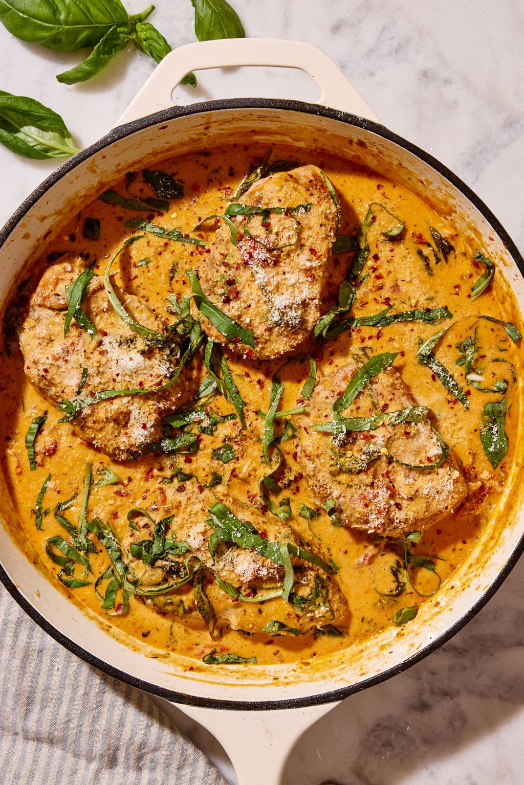 Overhead view of Marry Me Pork Chops nestled in a skillet of rich, creamy sun-dried tomato Parmesan sauce, garnished with fresh basil, grated Parmesan cheese, and red pepper flakes.