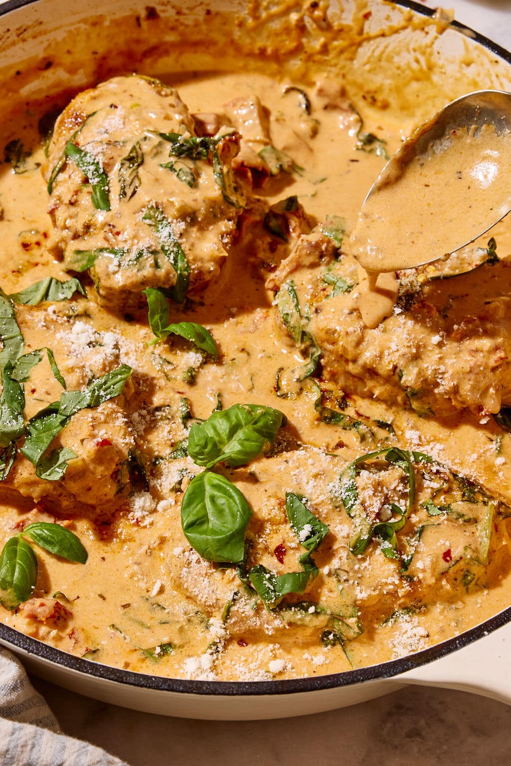 Close-up of Marry Me Pork Chops nestled in a skillet of rich, creamy sun-dried tomato Parmesan sauce with fresh basil and red pepper flakes, as a spoon drizzles extra sauce over the pork.