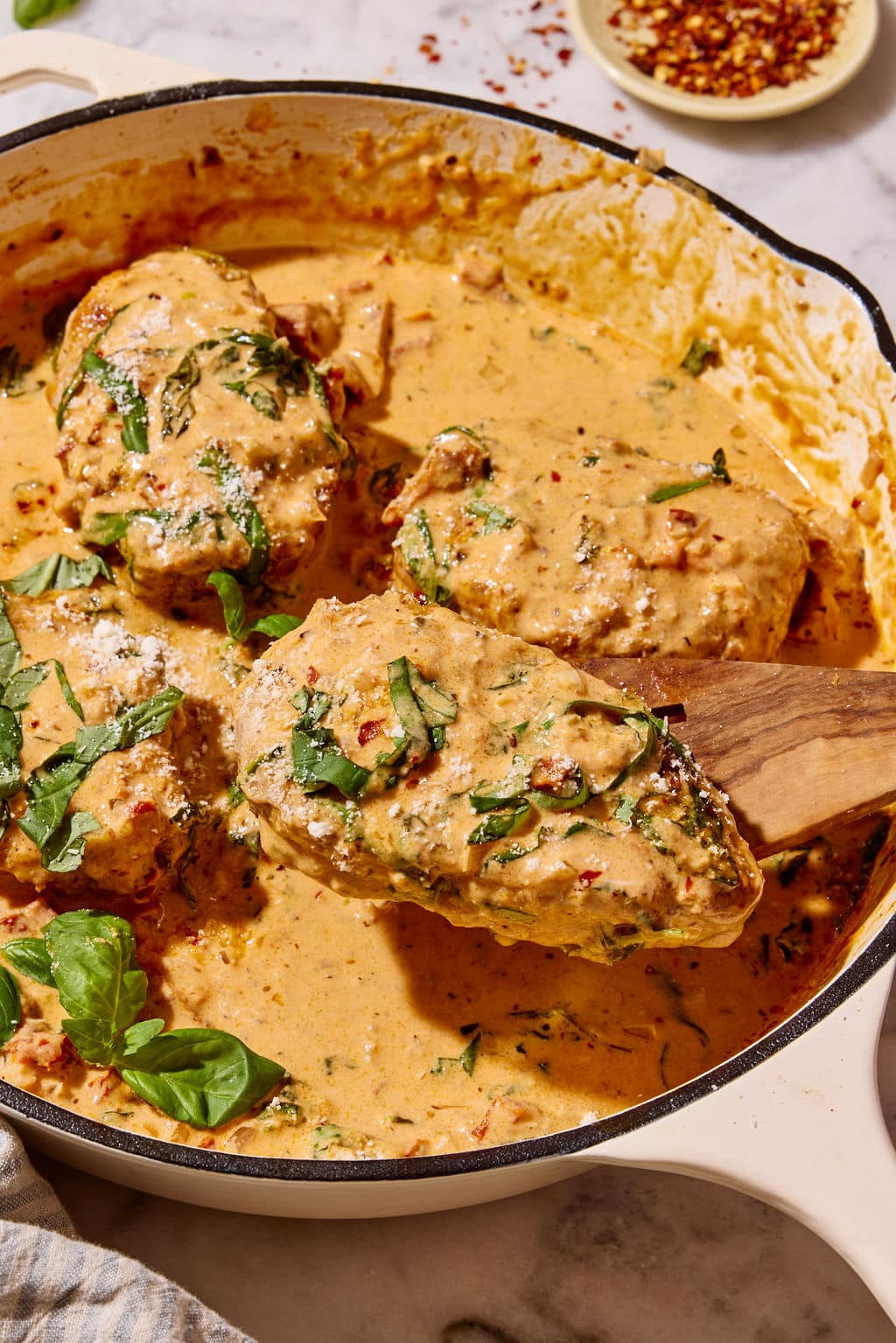 Golden-brown Marry Me Pork Chops coated in creamy sun-dried tomato Parmesan sauce, with one pork chop lifted from the skillet on a wooden spatula and garnished with fresh basil and grated Parmesan.
