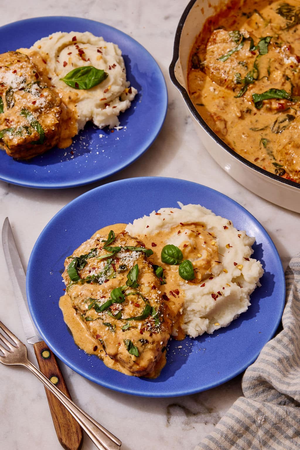 Marry Me Pork Chops served with creamy mashed potatoes on vibrant blue plates, topped with rich sun-dried tomato Parmesan sauce, fresh basil, and red pepper flakes, with the skillet of pork chops alongside.