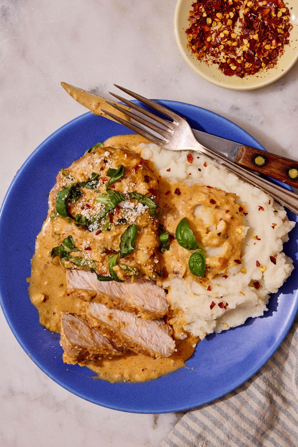 Overhead close-up of sliced Marry Me Pork Chops smothered in creamy sun-dried tomato Parmesan sauce, served alongside fluffy mashed potatoes on a blue plate and garnished with fresh basil and red pepper flakes.